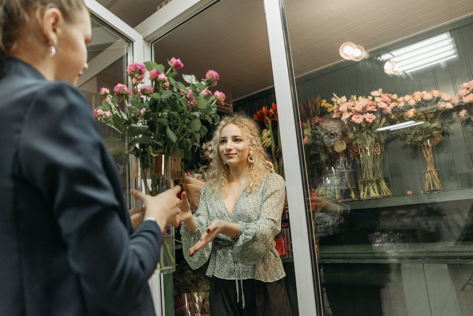 A woman with curly blonde hair, wearing a light floral blouse, is receiving a bouquet of vibrant pink roses and greenery from a flower shop employee inside a florist store. The bouquet is wrapped in b