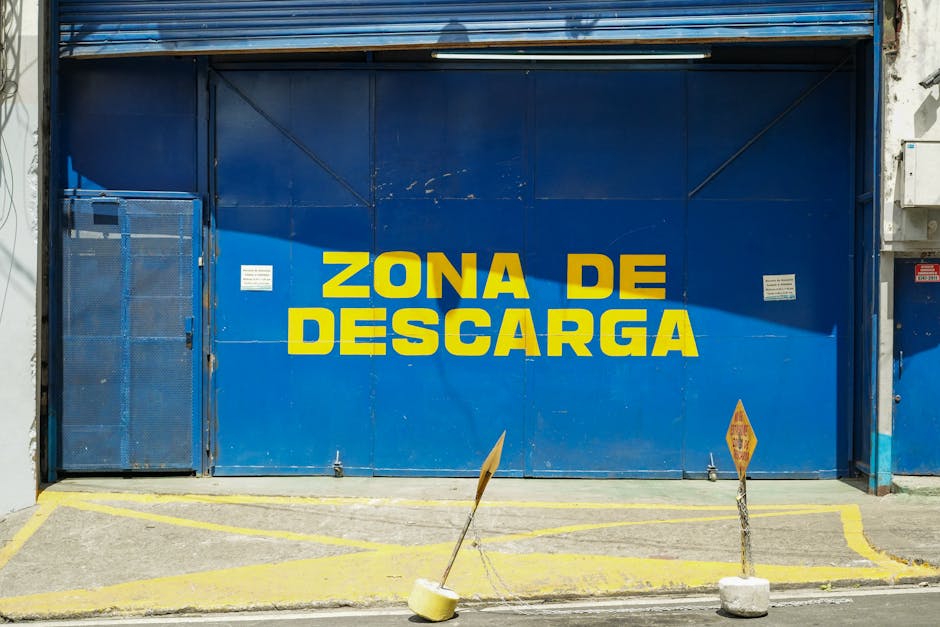 A large blue metal door with bold yellow letters displaying 'ZONA DE DESCARGA', indicating a delivery or unloading zone in Spanish. The door is set in an industrial building exterior, with a small blu