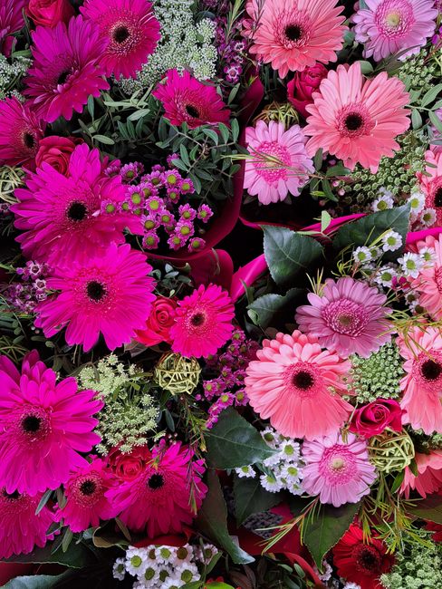 A vibrant floral arrangement featuring a mix of pink and magenta gerbera daisies, small white daisy-like flowers, and deep green foliage. The gerbera daisies vary in size and shade, with some petals f