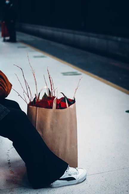 A brown paper shopping bag filled with fresh, vibrant flowers arranged with red blossoms and delicate sprigs of dried branches. The bouquet is positioned on a clean, light-colored airport or station f