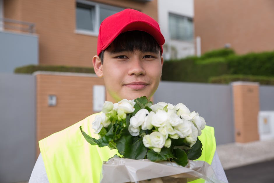 A young male flower courier wearing a red cap and neon yellow safety vest is delivering a bouquet of white roses wrapped in white paper. The roses have lush green leaves and are fresh with delicate, s