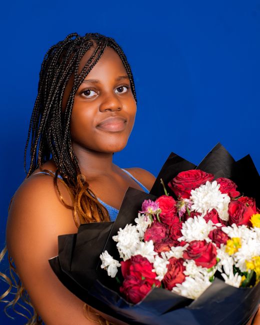 A young woman with braided hair holds a vibrant floral bouquet against a solid blue background. The bouquet features a mix of red roses, white chrysanthemums, and smaller yellow and pink blooms, arran