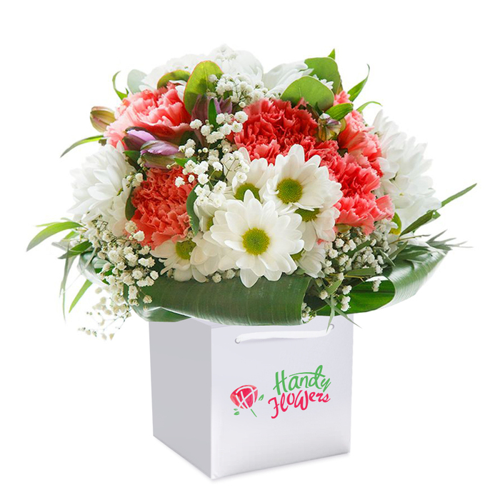 A radiant bouquet, masterfully arranged and thoughtfully presented in a spotless square white gift bag, anchors this striking image-its background a perfect canvas of blanc. The arrangement pulses with textural interest and vivid colour; at the forefront, ruffled carnation blossoms in coral, peach, and red hues unfurl their layered petals, carrying a gentle scent reminiscent of classic English blooms. Scattered throughout, fresh white daisy-like blooms, each with a zesty lime centre, introduce a dancing brightness to the overall palette. Wisps of white baby's breath weave dreamlike among the larger blooms, imparting a sense of airiness and movement familiar to those strolling the communal greenways of Marks Gate. Tucked discretely within, unopened buds-hinted with purple and pale pink-nod quietly to the promise of new beginnings. The base is framed in an elegant swirl of deep emerald and glossy green leaves, their broad, polished surfaces offering robust support and rich contrast to the flowerheads above. The bag's crisp, modern look, including sturdy white ribbon handles, ensures easy carrying-an inviting touch for flower delivery in this east London community. Every element is arranged for a generous, rounded finish, evoking freshness, optimism, and abundance. This visually lush composition represents the care, expertise, and local pride poured into every Mark's Gate bouquet, suitable for commemorating milestones or offering comfort in life's special moments.