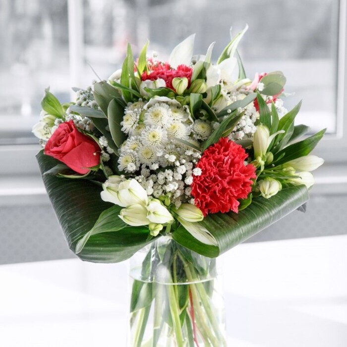 In this image, a beautifully composed bouquet of red and white flowers stands in a clear cylindrical glass vase, the stems neatly aligned and visible below the water, giving a sense of transparency and freshness. At the centre, deep red roses unfurl in velvety layers, their rich colour echoing the classic language of love. At the front, a generously ruffled red carnation draws the eye, its many frilled petals creating a soft, tactile texture that contrasts with the smoother rose petals. Around these passionate tones, a constellation of white blossoms provides balance: white alstroemeria with delicate speckles, rounded white chrysanthemums, and creamy accents lend an air of gentle calm. Several white lily buds remain closed, their elongated forms reaching upward and hinting at future blooms, like a promise that the arrangement will continue to evolve in the days after delivery. Wisps of baby's breath weave through the bouquet, their tiny white flowers floating like soft snow, adding lightness and volume without overwhelming the main blooms. The design is framed by luxuriant greenery: broad, glossy dark green leaves have been artfully curled and wrapped to form a polished collar at the base, while additional foliage in varying shades of green and eucalyptus-like textures weaves between the flowers, giving depth and structure. The bouquet sits on a spotless white surface against a softly blurred, bright background, as if placed near a sunlit window overlooking the quiet streets of Marks Gate. Prepared with the practiced hand of a local florist, it feels ideal for a dining table on Billet Road, a bedside in a Marks Gate flat, or as a same-day delivery surprise to celebrate an anniversary, birthday, or heartfelt thank-you in this close-knit community.